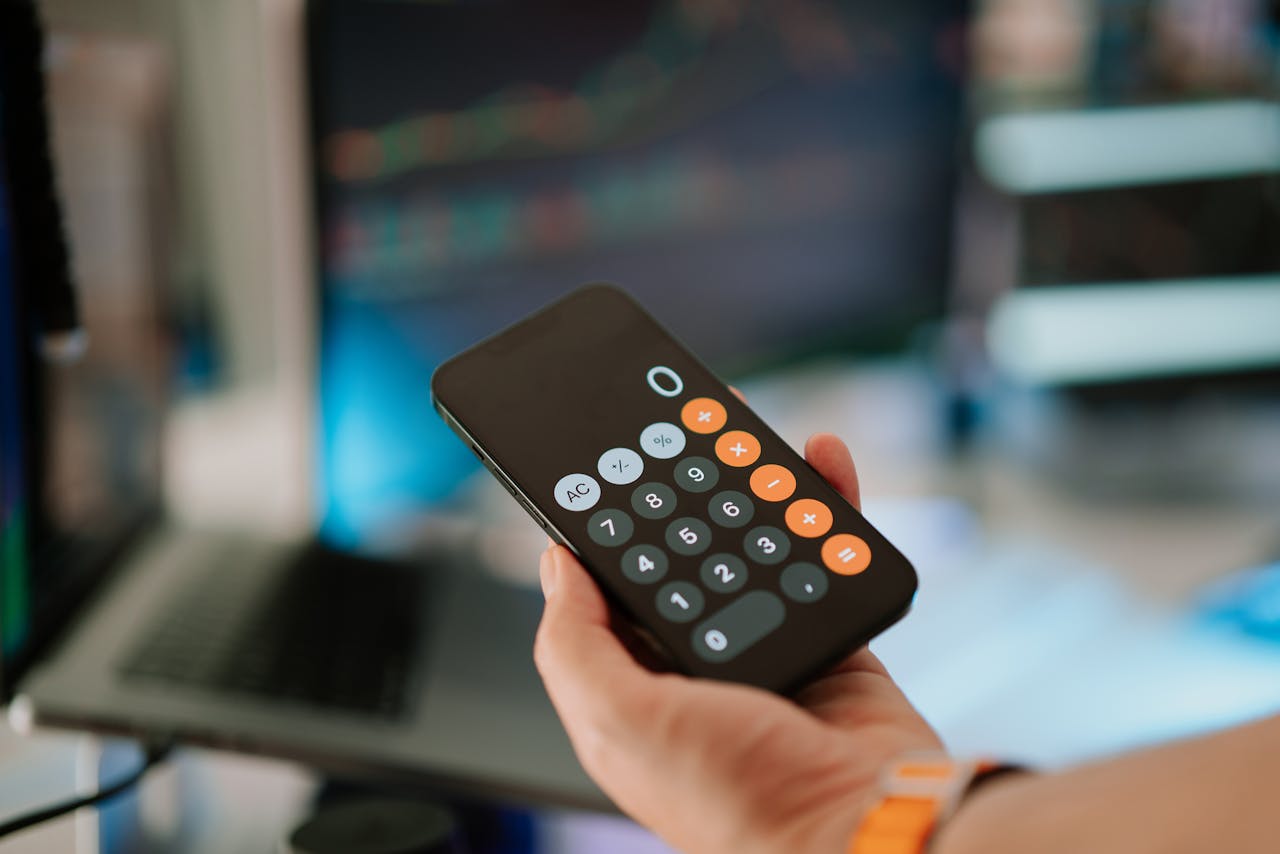 Close-up of hand holding smartphone calculator in office setting with financial data on screen.