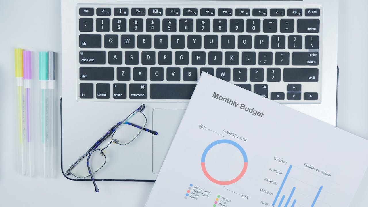 Aerial shot of a modern workspace with a laptop, budget report, and eyeglasses on a desk.